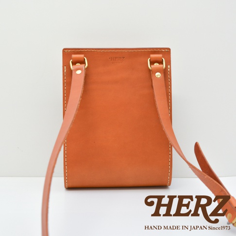 日本職人品牌 HERZ 手作皮革單肩包的背面視角，展現出厚實的 Latigo 硬皮革質感與暖棕色調。包身頂部有壓印 HERZ 與 MADE IN JAPAN 字樣，搭配黃銅色金屬環扣與堅固的皮革背帶，整體車縫線細節細膩，呈現簡約且經典的皮革工藝風格。