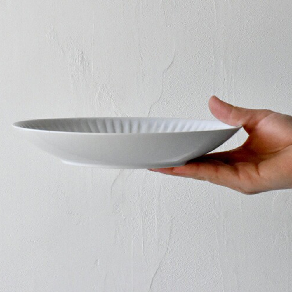 白色陶瓷主食盤，盤緣有放射狀的壓紋，由手托著，背景為淺灰色紋理牆面。