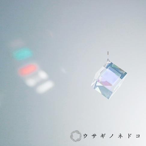 透明玻璃稜鏡立方塊吊飾，懸掛於細線上，折射出七彩光芒，左側牆壁上投射出紅、綠、白色的光斑。