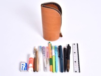 職人手作皮革直立式收納筆袋，呈現自然原色皮革，搭配金屬拉鍊。下方展示各式文具，包含橡皮擦、鉛筆、原子筆、水性筆及尺，色彩繽紛。