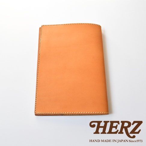 日本 HERZ 職人手作皮革筆記本書套，採用淺棕色 Latigo 軟皮革製成，表面呈現細膩溫潤的皮質紋理，邊緣以淺色縫線細緻包邊，呈現簡約耐用的經典書套設計，下方附有品牌標誌與創立年份說明。