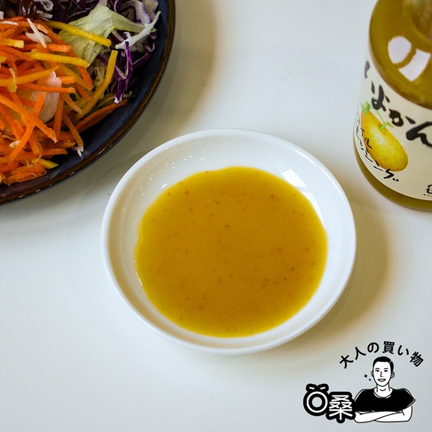 一碗盛有黃色柑橘風味沙拉醬的白色圓形小碟，旁邊是一瓶黃色瓶蓋的玻璃瓶裝沙拉醬，瓶身上有日文標籤和柑橘圖案。左側有一盤混合蔬菜沙拉，包含切絲的紅甘藍、生菜和胡蘿蔔。