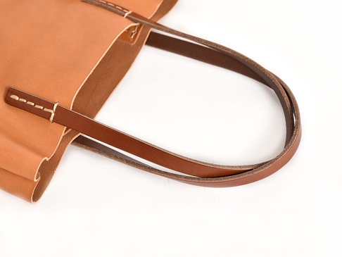 職人手作皮革直式軟皮托特包，採用淺棕色皮革製成，配有較深的棕色皮革提把，手工縫線細緻，展現簡約質感。