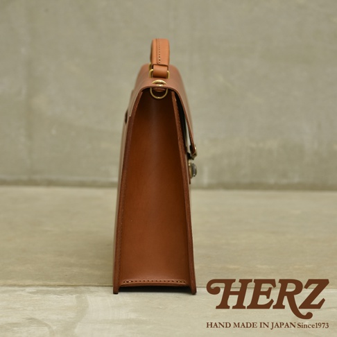 職人手作皮革Organ兩用商務公事包，呈現經典的棕褐色調，以高品質皮革製成，側邊視角展現其簡潔俐落的線條與精緻的縫線細節，配有金色金屬鎖扣和皮革提把，整體散發出低調的商務質感。