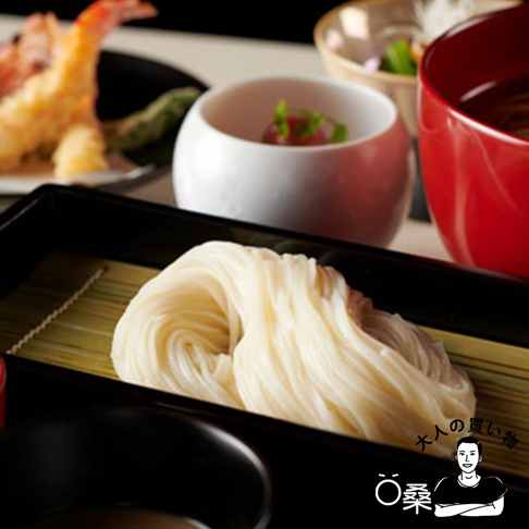 一盤白色的無選別稻庭烏龍麵，麵條捲成一團，放置於竹墊上的黑色長方形盤中，背景有紅色湯碗、白色小碗及炸蝦等日式餐點。