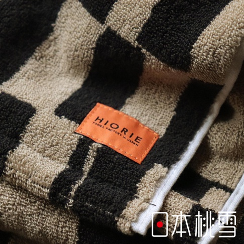 日本桃雪戶外格紋棉柔2WAY連帽浴巾，織有焦糖色和黑色的棋盤格圖案，標籤上印有HIORIE TOWEL FACTORY in JAPAN字樣。