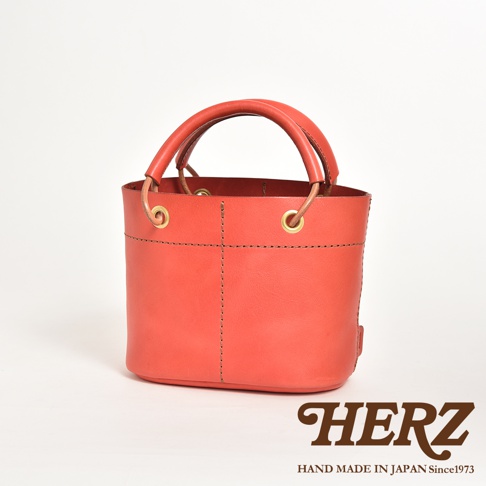 珊瑚橘色皮革製成的手提午餐袋，設有加厚提把與大開口，袋身有縫線細節與金色金屬孔眼，底部平整。