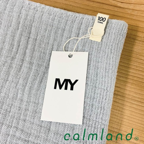 淺灰色無撚紗質長毛巾，表面織紋細緻，帶有「MY」品牌吊牌及「100 TOWEL」小標籤，材質觸感柔軟。
