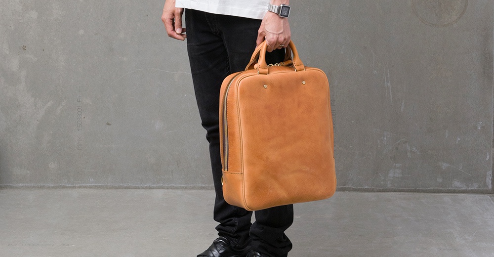 一名男子提著一個棕褐色皮革後背包，背包具有簡潔的方形設計和頂部提把。