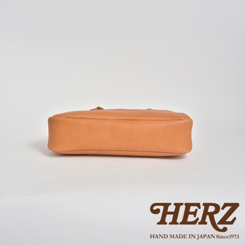 一張展示日本HERZ職人手作皮革肩背托特包底部的特寫照片，以純淨的淺灰色背景為襯，呈現出皮革自然的原色質感與平整的包底結構，右下角標註有HERZ品牌名稱及日本手工製作年份資訊。