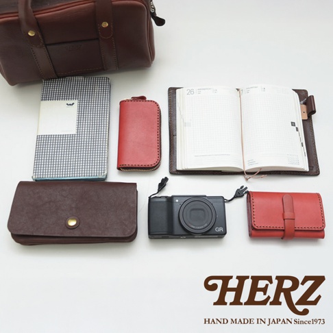 日本HERZ手工皮革製品，包含一個咖啡色兩用迷你波士頓包、一個棕色長夾、一本紅色鑰匙包、一本黑色數位相機、一本灰格子筆記本和一本日誌。