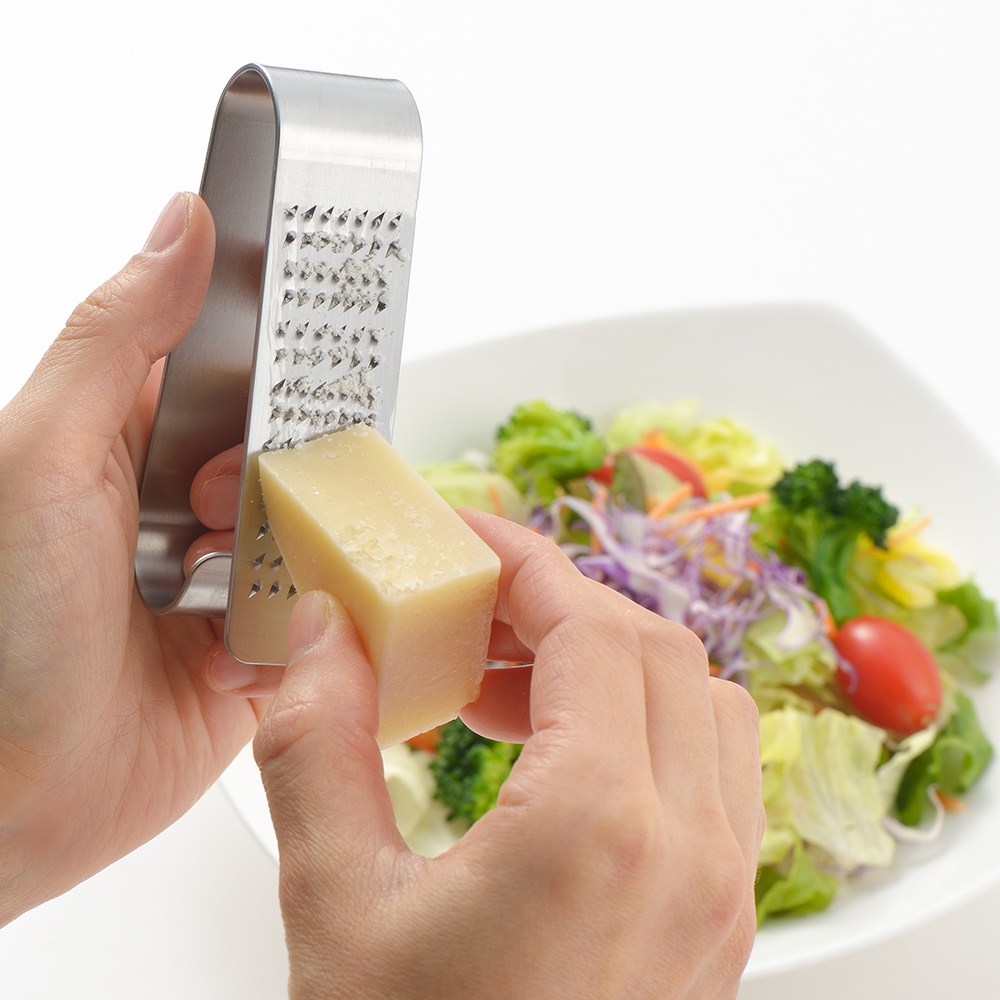 日製立式磨泥器，由不鏽鋼製成，設有細密磨孔，使用者正用手握持磨泥器，將一塊淺黃色起司磨碎，背景為一碗色彩豐富的沙拉。