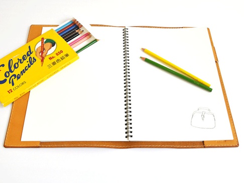 淺棕色皮革A5筆記本書套，攤開展示內頁，側邊放有黃色與綠色鉛筆，上方擺放一盒彩色鉛筆。