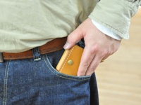 職人手作皮革箱型收納零錢包 (KK-23)，採用天然皮革製作，呈現溫潤的焦糖色調，表面有精緻縫線。零錢包以箱型設計，正面設有金屬按扣，由手部放入牛仔褲口袋中。