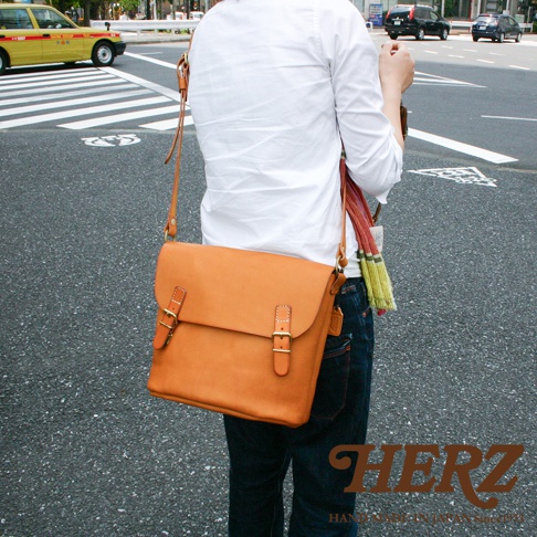 模式B： 日本HERZ職人手作橘色皮革雙扣旅行郵差包，背在身穿白襯衫、藍色牛仔褲的人身上，包包細節與品牌標誌清晰可見。