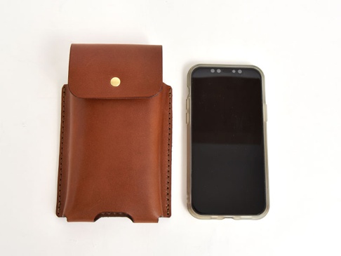 一個棕色皮革手機收納包，帶有金色按鈕封口，旁邊是一個裝有透明保護殼的黑色智慧型手機。