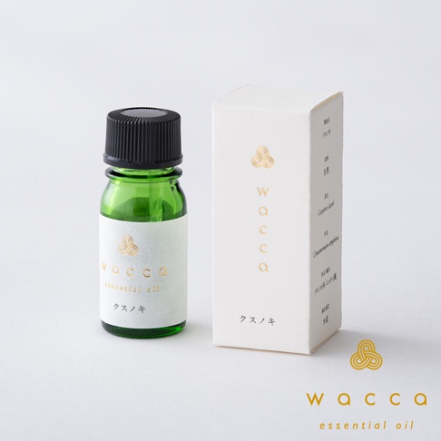 綠色玻璃瓶裝有佐賀楠木精油，瓶身貼有白色標籤，印有金色「wacca essential oil」字樣和楠木圖案。旁邊有一個白色紙盒，同樣印有金色「wacca」標誌和楠木名稱。