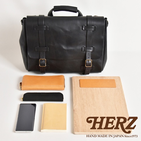 黑色皮革復古郵差包，搭配兩條皮質束帶及金屬釦環。下方擺放一個棕色皮革長夾、一個黑色筆袋、一本米色筆記本、一支手機及一個木質夾板，夾板上有皮革飾片及品牌標誌。