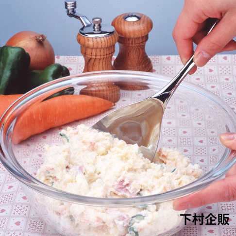 一只不鏽鋼料理分食匙，匙面寬大，用於舀取玻璃碗中的馬鈴薯沙拉。背景有胡蘿蔔、洋蔥、青椒和木質鹽罐胡椒罐。