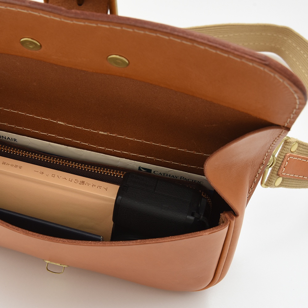 職人手作皮革單扣經典斜背包，呈現原色皮革材質，內部可見書籍、相機等物品，搭配帆布背帶與金屬扣環。