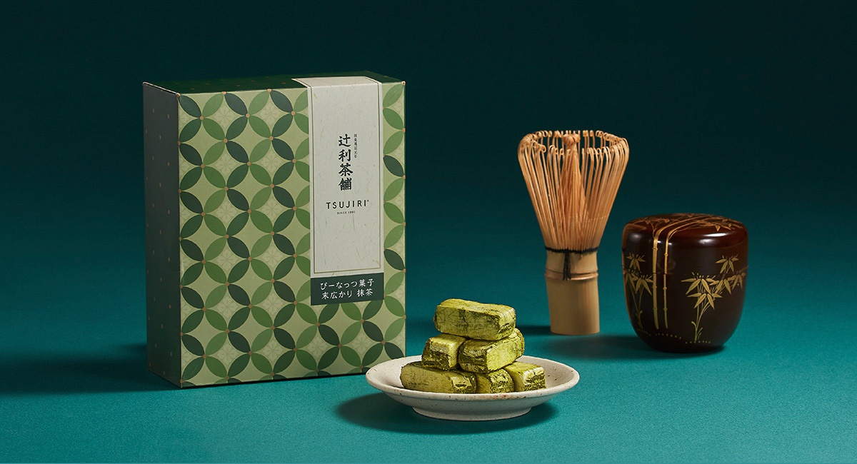 辻利茶舖的抹茶花生酥禮盒，放置於深綠色背景中，左側為印有綠色幾何圖案與品牌標誌的長方形禮盒，中心盤子上堆疊數塊抹茶粉包裹的酥脆花生糖，右側則擺放竹製茶筅與繪有竹紋的黑色茶罐，整體呈現出沈穩優雅的日式茶點意象。