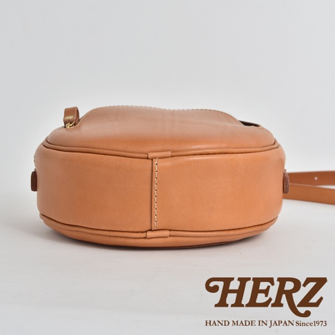 淺棕色皮革小圓包，側面視角，包身圓潤飽滿，縫線精緻，搭配皮質提把與肩帶。