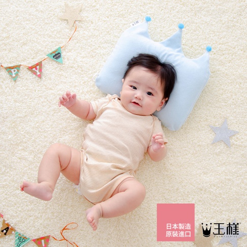 嬰兒躺在淺色絨毛地毯上，頭枕著一個淺藍色皇冠造型的枕頭，枕頭上有兩個藍色絨球裝飾。嬰兒穿著一件米白色條紋連身衣。背景有掛著「PY」字樣的彩色三角旗幟和一個銀色星星裝飾。