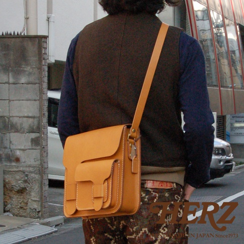 職人手作經典厚實單肩包，採用黃褐色皮革製成，設計簡約，帶有明顯的縫線細節，並配有可調節的長肩帶。