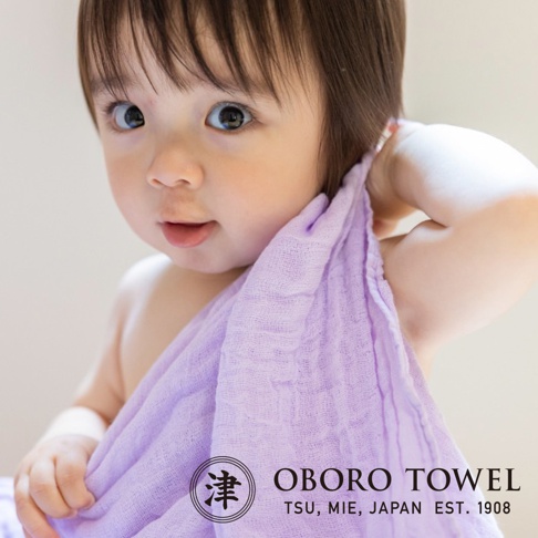 一名嬰兒被一條淡紫色三層紗純棉裹身毯包裹著，露出可愛的表情。