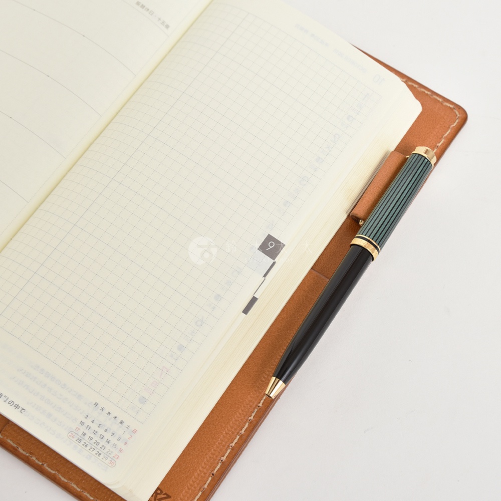 職人手作皮革旅行記事本書套，展示一本攤開的筆記本，內頁為方格網線，並有一支黑色筆身、綠色筆蓋的鋼筆置於書套內。