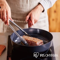 黑色不沾平底鍋中，一塊牛排正被夾子翻動。平底鍋帶有木紋把手，置於電磁爐上。