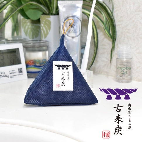 深藍色網狀材質的立體三角調濕木炭，頂部有金色繩圈，正面貼有白色標籤，上面有日文文字和一個黑色圖案。