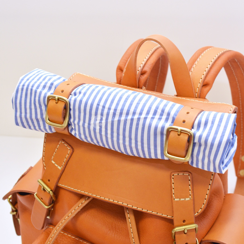 職人手作皮革復古風旅行後背包，主體為棕褐色皮革，頂部捲筒處包裹著藍白條紋布料，以皮革束帶與黃銅釦環固定。
