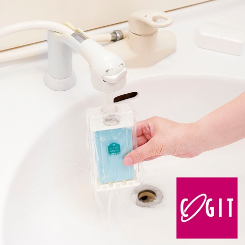 透明塑膠盒裝著淺藍色洗衣皂，下方有白色塑膠刷頭，被一隻手持著，置於水龍頭下方，水流正沖在盒子上。