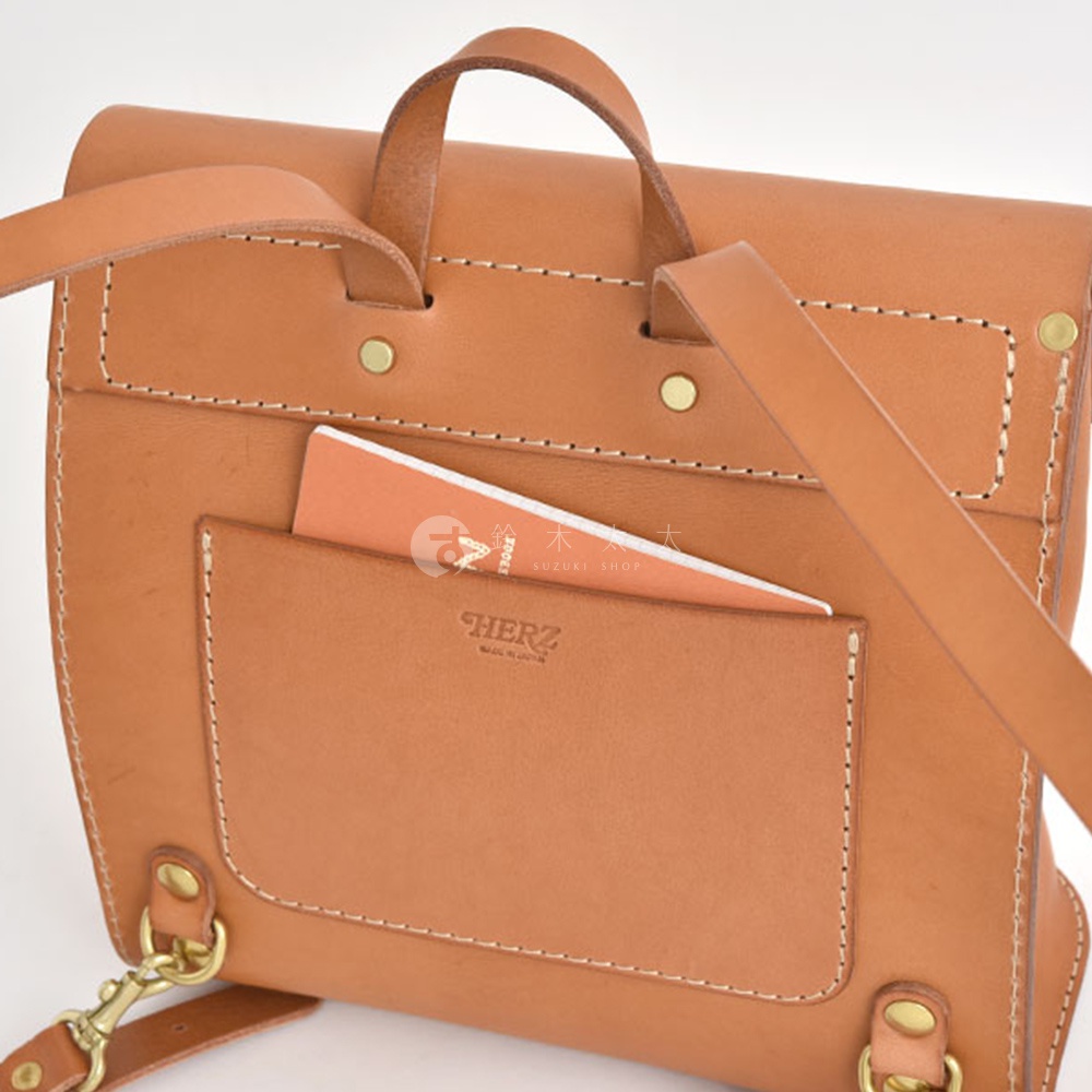 職人手作皮革翻蓋迷你後背包，主體為淺棕色皮革，縫線清晰，設有外插袋，袋中露出橘色筆記本，品牌「HERZ」壓印於外袋上。