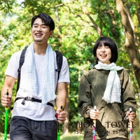 一對男女在戶外健行，皆配戴淺藍色三層紗純棉掛脖毛巾。男士穿著白色T恤，女士穿著軍綠色外套。兩人皆持登山杖，背景為綠意盎然的樹林。