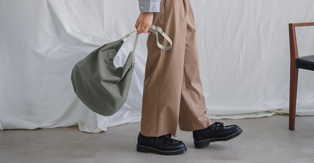 身穿卡其色寬褲與黑色樂福鞋，手提淺綠色皺褶感肩背托特包，背景為白色布幔與木質椅