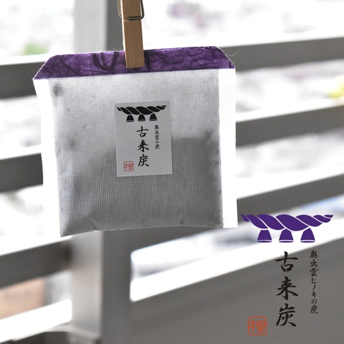 一個夾著木夾懸掛的白色網狀小袋，袋口飾有紫色紙張，袋上貼有標籤，標籤上有黑色漢字「古來炭」及其他日文文字，袋內可見黑色木炭顆粒。