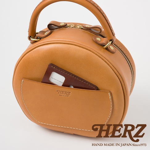 HERZ 品牌標誌與 Made in Japan 字樣，以及一顆卡片夾在棕色皮革包的口袋裡，包包材質為焦糖色皮革。