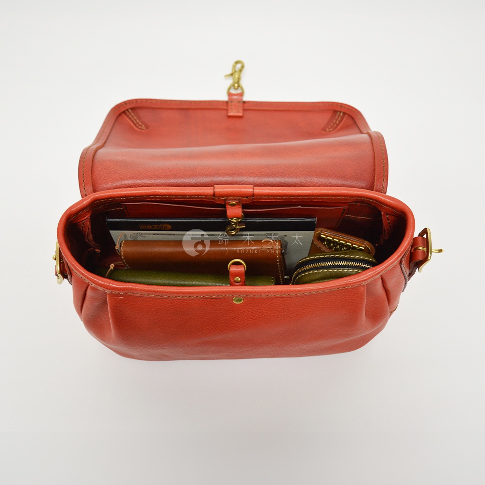 職人手作皮革復古翻蓋郵差包，採用橘紅色皮革製成，包身飽滿，內有書籍、錢包、零錢包等物品，搭配金色金屬釦環和側背帶。
