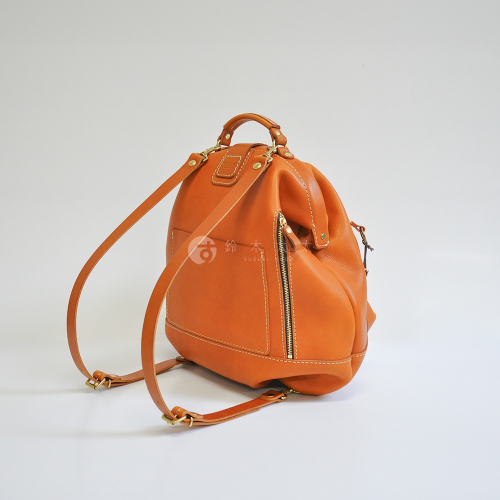 職人手作皮革輕旅行後背包 R-144，採用橘棕色皮革，設計簡潔，設有拉鍊側袋及可調節肩帶。