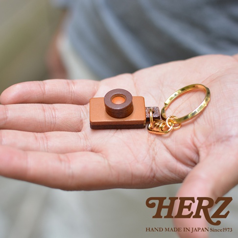 職人手作皮革相機造型鑰匙圈，主體為淺棕色皮革，搭配深棕色鏡頭細節。鑰匙圈附有金色金屬環和扣環，整體設計精緻小巧。