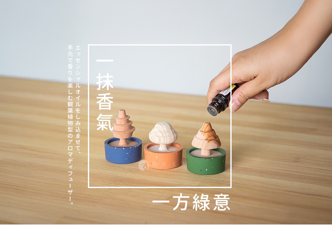 一隻手正將精油滴在木質擴香樹上，桌面上擺放著三個裝飾性擴香台，底座分別為藍色、橘色與綠色，上方搭配不同形狀的木質樹木造型。圖片兩側配有中文文字，說明這是一款可藉由滴入精油享受芳香的觀葉植物造型擴香器，旨在營造室內的一抹香氣與一方綠意。