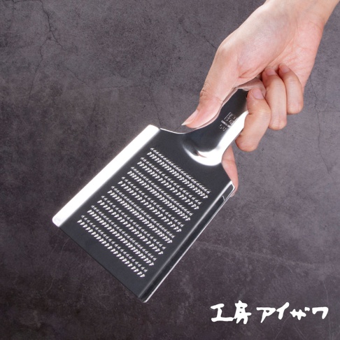 一只手正握著一把銀色不鏽鋼製的食材磨泥器，磨泥器表面佈滿細小的磨孔，手柄處有品牌標誌。