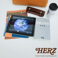 皮革軟式杜勒斯包，包含一個平板電腦、一本筆記本、一本教科書和一個保溫杯，產品以棕色皮革為主，搭配金屬拉鍊和鉚釘。