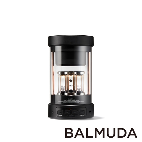 The BALMUDA wireless speaker is cylindrical in shape with a black top and base. A clear glass cylinder encloses three glowing vacuum tubes with copper accents. The base features control buttons for po