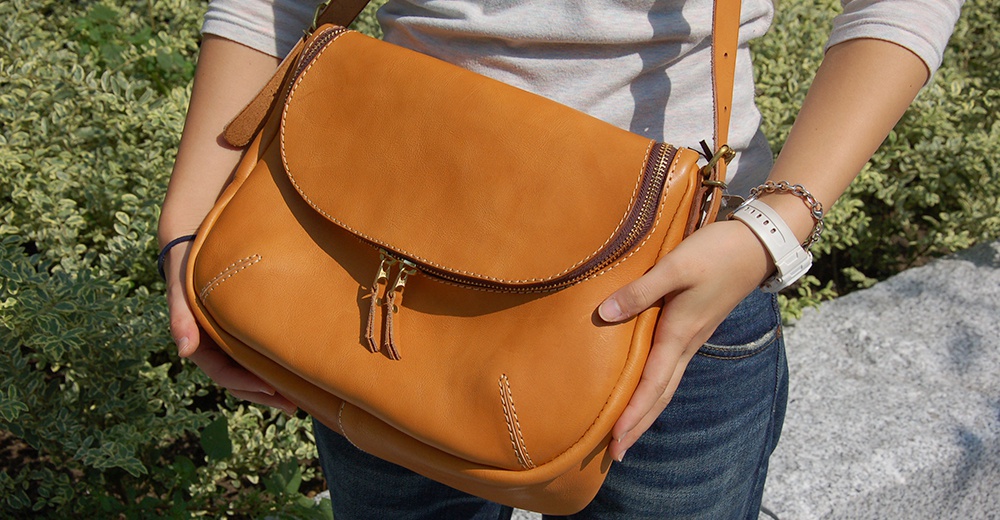 日本HERZ職人手作Stare皮革大容量馬鞍包CK-150，駝色，側面拉鍊與雙拉頭設計，搭配牛仔褲與手錶，背景為綠色植物與石頭。