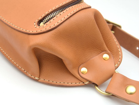 職人手作皮革拉鍊旅行斜背包，採用天然皮革材質，呈現溫潤的棕褐色。包身設有黃銅色拉鍊，邊緣以米白色縫線精緻縫合。包袋一側有兩個黃銅色鉚釘固定，並連接一個金屬環，風格簡約實用。