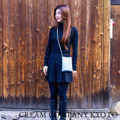 淺藍色皮革手提隨身手機包，搭配白色細肩帶，由女性模特兒展示，身穿黑色高領毛衣、黑色百褶裙和黑色長靴，背景為深棕色木紋牆。