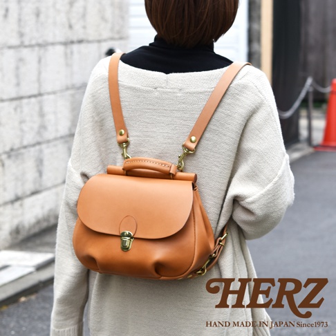 一位女性背著日本HERZ職人手作皮革後背包，該包款呈現溫潤的焦糖棕色，具備獨特的屋瓦造型翻蓋設計與金屬扣環，搭配黃銅色五金細節，整體風格簡約且充滿手作質感。